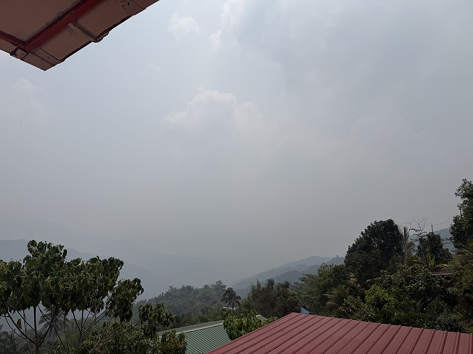 Pemandangan sebuah kawasan kampung diselubungi jerebu kelabu tebal, dengan bumbung rumah berdiri di tengah pokok hijau sementara bukit di latar belakang kabur dalam jerebu